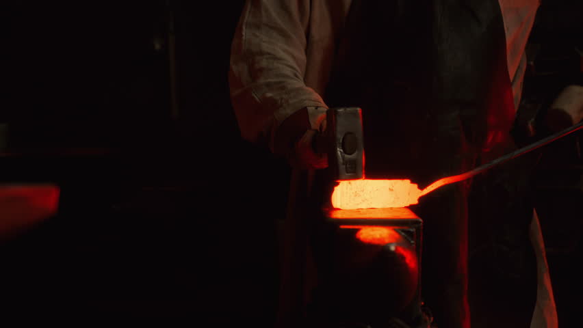 A blacksmith uses a hammer to shape a piece of red-hot metal in a forge on an anvil, creating a future product, sparks fly from blows. The process takes place in the darkness of the forge.