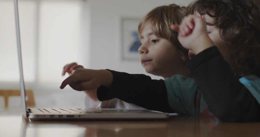 Two small boys concentrate on a laptop screen, with one child pointing at something interesting, sharing a moment of discovery and learning in their living room.