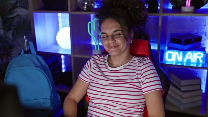 A happy young woman with curly hair sits crossed-armed in a modern gaming room at night.