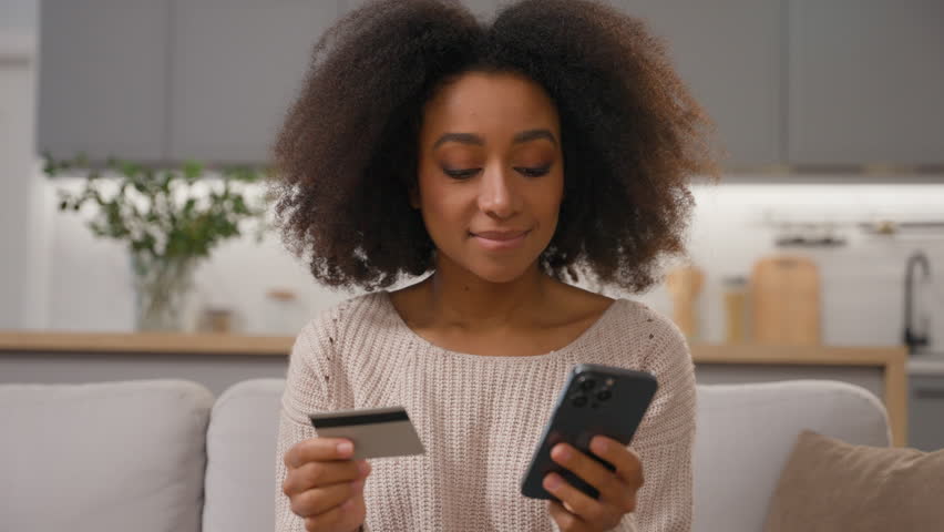 Happy smiling African American woman hold mobile phone and credit card shopping online order delivery with smartphone buying pay money ethnic girl female shopper use digital banking at home kitchen Royalty-Free Stock Footage #3486843981