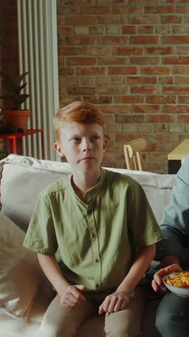 A man and two boys are sitting on the couch and eating snacks while watching TV at home.
