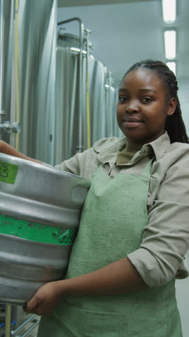 Medium vertical portrait shot of happy young African American woman in apron, with braided hair, standing beside steel vats in brewery, holding beer keg and looking at camera and smiling