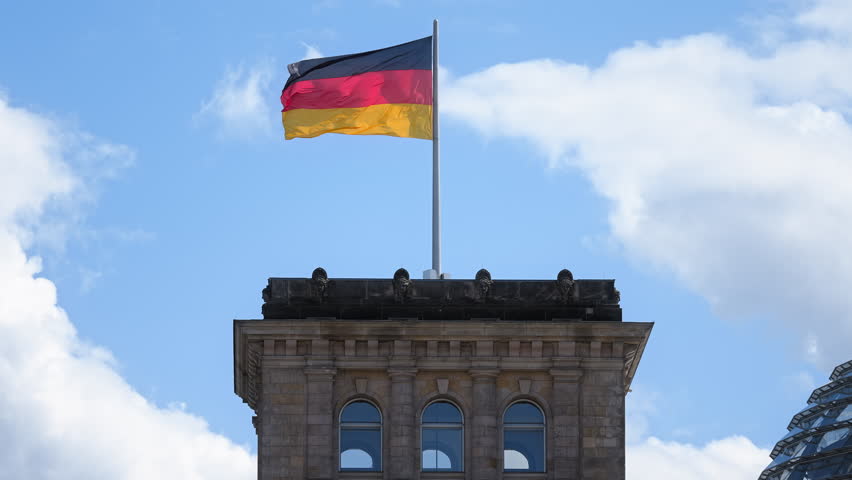 Germany flags wave against blue sky. Deutschland government symbol close up. City street background. Patriotism proud concept. National german silk flag on building roof top. Berlin sign fluttering.