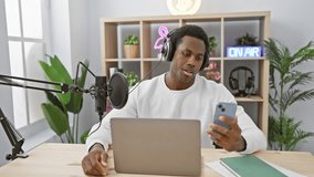 Young black man podcasting from a studio with headphones, smartphone, laptop, microphone, and 'on air' sign.