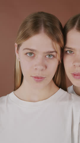 PAN vertical portrait of beautiful young twin sisters in white t-shirts standing together isolated on brown background and looking at camera