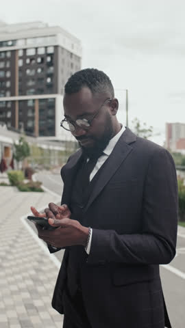 Vertical tracking of African-American businessman in suit and glasses standing on street and using mobile phone