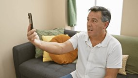 Middle-aged man in white shirt holding smartphone for a video call in a modern living room setting.