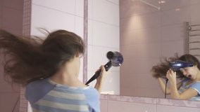 Beautiful happy girl teenager dries hair with a hair dryer and sings and dances in front of a mirror in the bathroom stock footage video