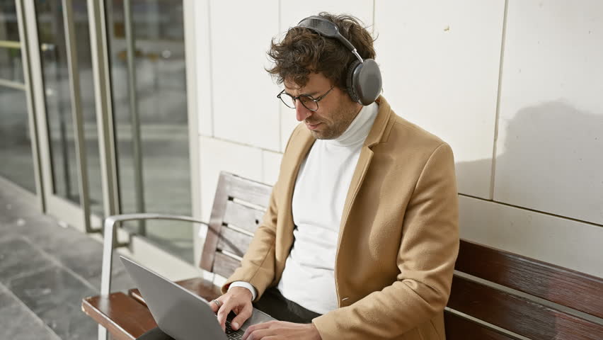 Handsome hispanic man with headphones using laptop on city bench outdoors