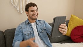Smiling man using tablet for video call at home, sitting on couch in casual clothes.