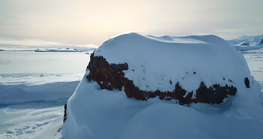 Fly over frozen Antarctic snow covered land in sunset. Untouched wilderness of South Pole. Desert white landscape, snow and ice aerial drone shot. Mountains in background. Nature polar conservation