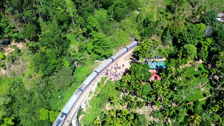 Aerial drone scenic view of train with locomotive carriages travelling along railway line entering into tunnel with people standing near Ella Sri Lanka Asia