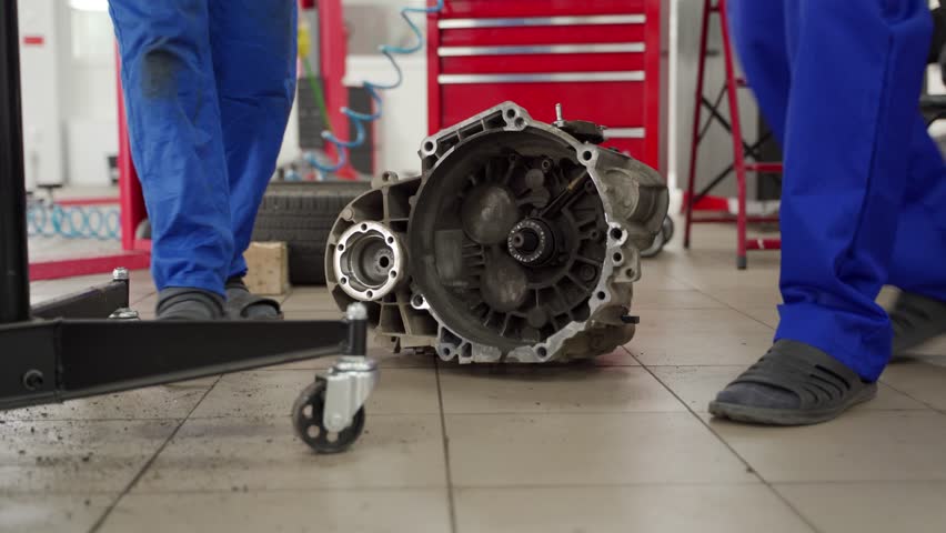 Auto mechanics in uniform remove car gearbox at repair shop. Teamwork in vehicle maintenance, manual transmission system servicing. Hands-on training in automotive tech, precise machinery work.