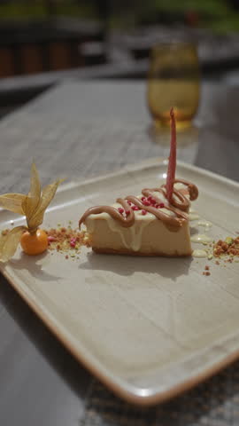 A delectable cheesecake garnished with a physalis and vibrant edible decorations on a ceramic plate with a blurred glass in the background.