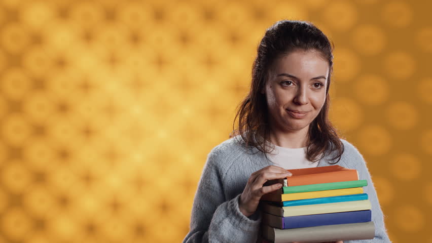 Portrait of smiling woman pointing towards empty copy spaces while holding pile of books. Radiant person with stack of novels in arms promoting reading, showing messages, studio background, camera A