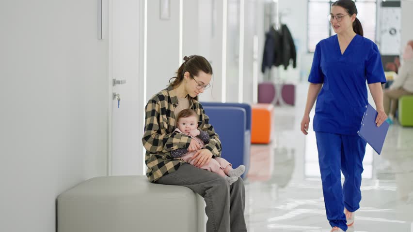 A young brunette girl the mother of a little baby girl sits in the corridor of a modern clinic and waits for her turn in the office of a pediatrician doctor. While a brunette girl doctor in a blue Royalty-Free Stock Footage #3487328889