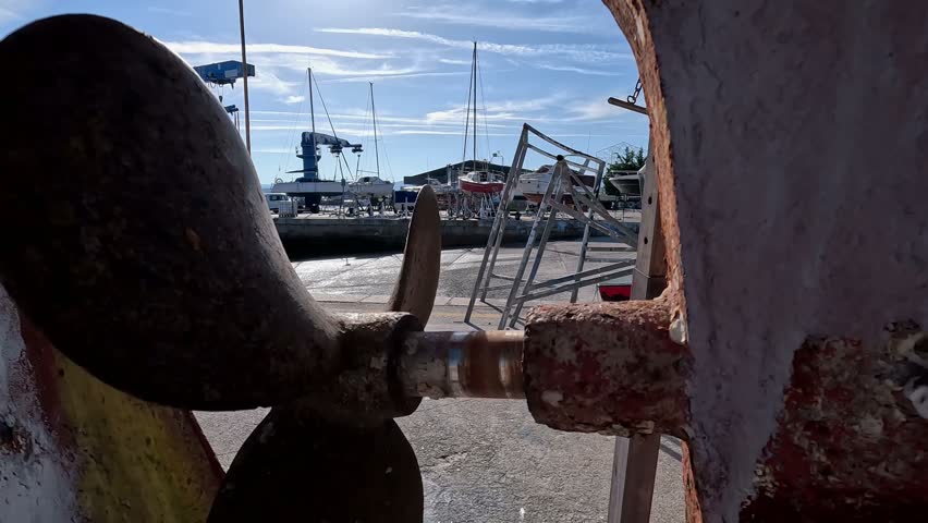 The rusty engine and rudder propeller of the old fishing boat in the shipyard for restoration with the harbor cranes in the background, close-up shot traveling backwards. Portonovo, Galicia, Spain