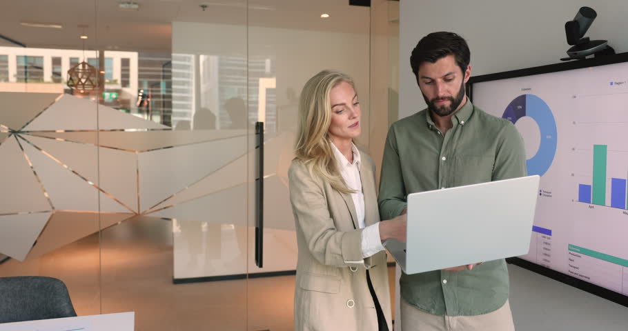 Two business partners stand near screen displaying charts and graphs, looking at laptop screen talking, reviewing data, trends, discussing insights, make decision, planning future marketing campaigns Royalty-Free Stock Footage #3487401783
