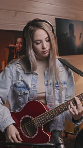 Vertical medium slowmo portrait of attractive young female guitarist in vintage oversize blue denim jacket looking at camera while playing acoustic guitar in old-school music studio Royalty-Free Stock Footage #3487523663