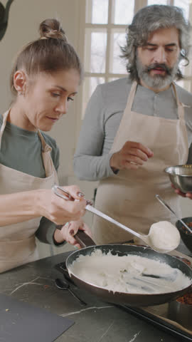 Slow-motion vertical medium shot of man and woman cooking lasagna during cooking class, woman pouring some Bechamel sauce on top of dish