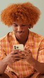 Vertical video, Smiling guy with curly red hair, wearing an orange shirt, uses mobile phone to text sitting in living room