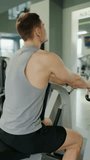Young Man Focused on Building Muscle at a Modern Gym. A young male athlete using a rowing machine at the gym, focused on his fitness goals, illustrating determination and strength. Vertical video.