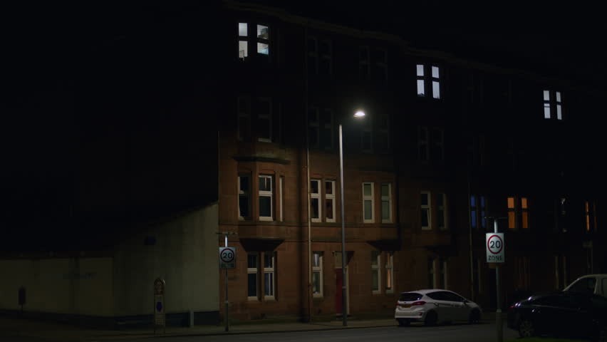 Street light flashes above speed limit sign as tall lamp illuminates road below apartment buildings at night. Faulty lamp