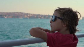 A boy wearing sunglasses, his gaze absorbed by the sweeping vistas of Istanbul as the ferry cuts through the waves, a backdrop of city life unfolding behind him. Slowmotion video.
