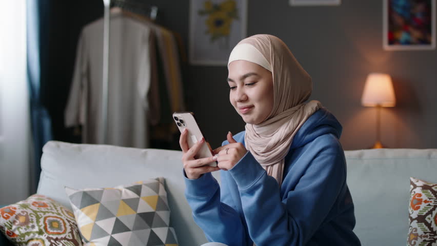 Muslim woman using phone, laughs, smiles. Muslim young teenage woman scrolls through videos, news on phone in social networks, browses web pages, online applications, uses smartphone and Internet. Royalty-Free Stock Footage #3487935707