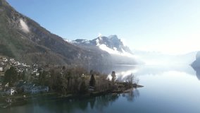 Bright drone clip with lens flare showing misty snow topped mountains next to calm lake in Switzerland