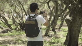 Young traveler walking through a forest and taking photos and recording videos of the landscape with his cell phone, enjoying the outdoors