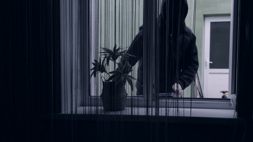 A thief or bandit in a mask breaks open a window with a crowbar and breaks into the house.