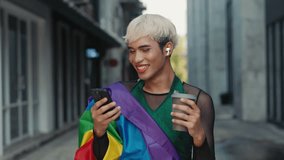 Smiling Man Walking Down the Street in LGBT Flag and Using Phone and Carrying Eco Cup with Coffee. Queer Man Outside on Coffee Break Walking and Watching Funny Videos. LGBT and Technology Concept - Powered by Shutterstock - Get 15% off with code: PIKWIZARD15