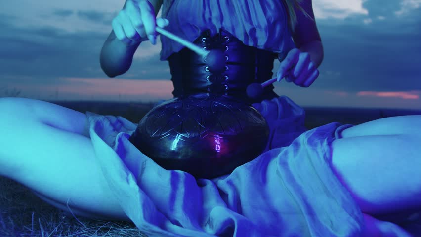 Young performer plays the hapi drum in blue neon light, against the backdrop of an inspiring and meditative evening sky. She are sitting on the grass, in the lotus position, surrounded by nature