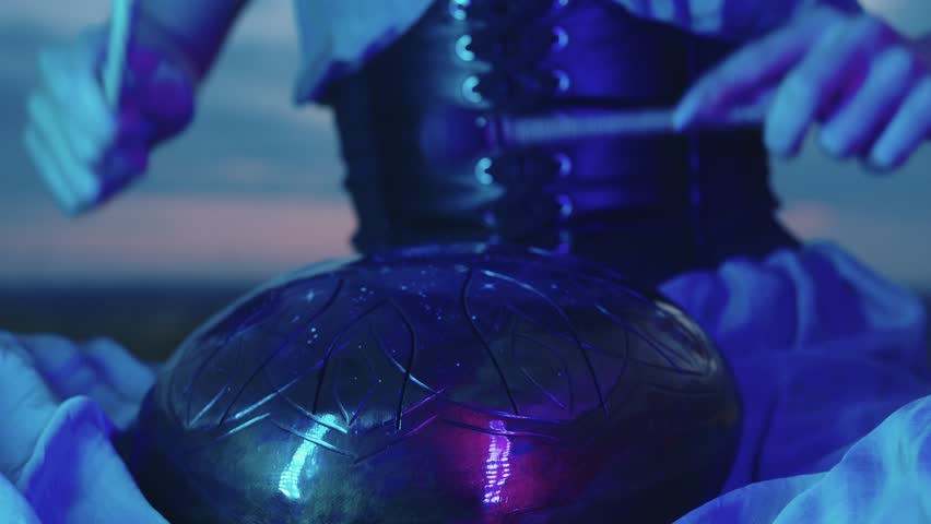 Girl is playing a steel-tongue drum while wearing a dress and a corset. She is meditating while sitting on the grass, with the evening blue sky as a backdrop. Close up. 4k