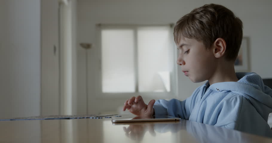 A young boy with blond hair is focused on using a digital tablet while sitting at a wooden table, in a well-lit, spacious living room during the day.