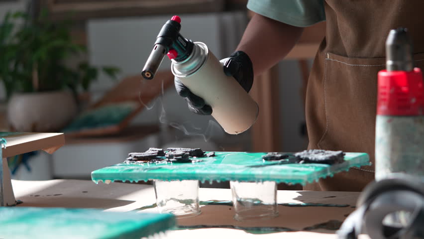 Woman artist using manual gas burner works with fire on wooden boards with epoxy resin. Pour art. Trendy modern hobby. Creating beautiful sea waves, landscapes with acrylic colours and epoxy. Art deco