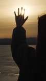 A praying woman stretches out her hand to the sun against the sunset, asking for God's blessing. Vertical video, shorts.