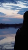 The silhouette of a girl raising an Orthodox cross to the sky at dusk. A mirror image of the cloudy sky on the surface of the water. The concept of religion and faith in God. Vertical video, shorts.