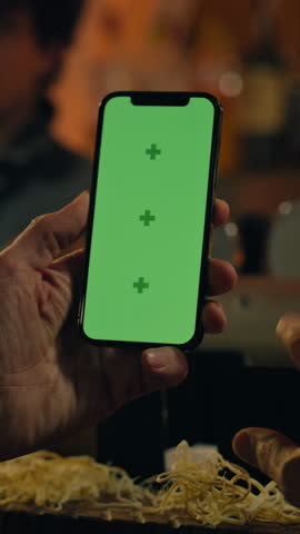 Vertical shot of Mature man uses mobile phone with green screen, bets on football match online or surfs the Internet. Sports fan checks bookmaker ratings, talks with bartender sitting at bar counter