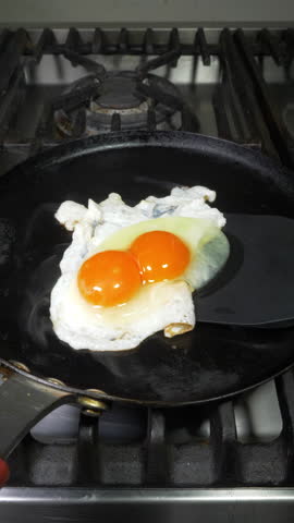 Vertical video - Closeup of a large double yolk hen’s egg, being turned over with a spatula as it fries in a steaming hot pan, on a kitchen stove. Royalty-Free Stock Footage #3488352479