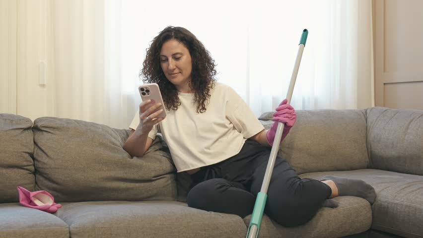 Tired curly woman in rubber gloves checking smartphone, sitting on sofa after cleaning apartment