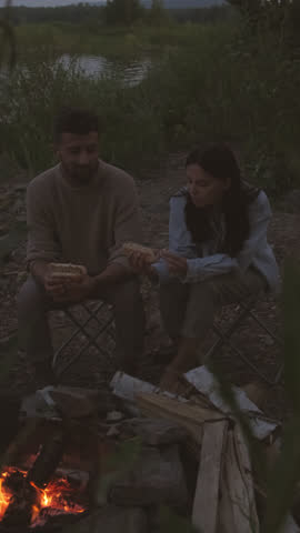 Vertical of bearded Biracial man sitting with dark-haired girlfriend on lakeshore by campfire on summer evening, eating hot dogs, talking and enjoying nature