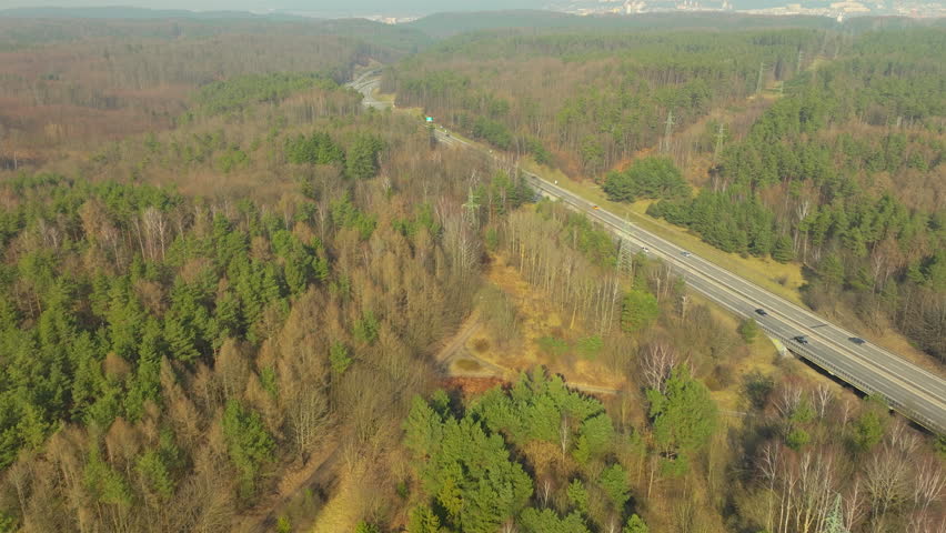 aerial view capturing a stretch of highway meandering through a forested landscape. Trees display a mix of evergreen and deciduous varieties, with seasonal change, in spring