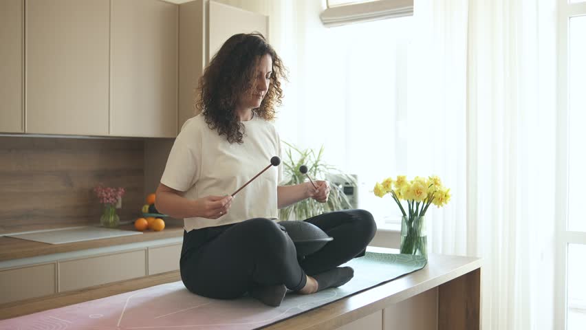 Relaxed curly brunette sitting in lotus pose on the kitchen table and playing tank drum