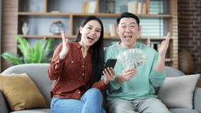 Happy asian married couple holding a fan of cash money dollar bills and phone while sitting on sofa at home looking at camera. They are satisfied with earning money using an application on smartphones - Powered by Shutterstock - Get 15% off with code: PIKWIZARD15