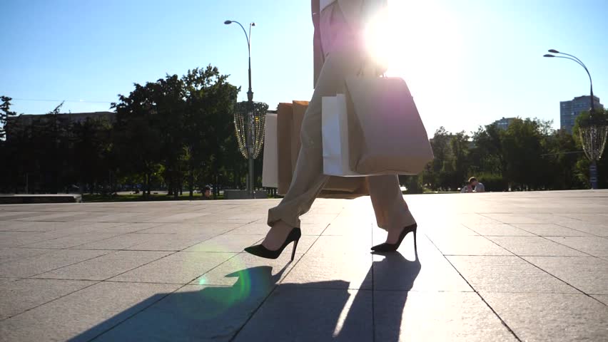Fashionable woman carries paper packets going along urban street. Young female customer holds shopping bags walks at square after purchases. Concept of leisure at sales and discounts time. Slow motion