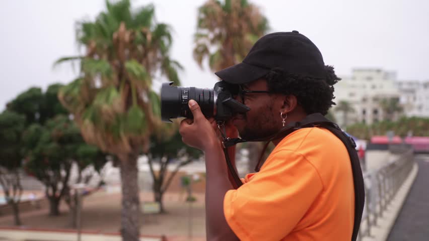 Stylish Black man with orange t-shirt, cap and cross earring holds camera near palm trees in Cape Town, South Africa
