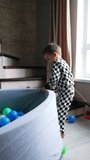 Two-year baby boy pulls the dry pool with balls by the room. Kid raises on the stair and drops himself into the basin. Vertical video.