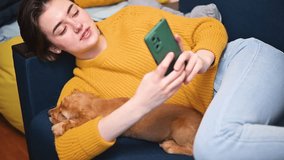 Cute beautiful young woman lying on the sofa with her pet puppy and using a smartphone, talking on a video call on the phone